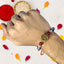 Hand wearing a beaded rakhi with a decorative charm, surrounded by colorful petals on a white background.