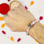 Hand wearing a beaded rakhi with a butterfly charm, surrounded by colorful petals on a white background.