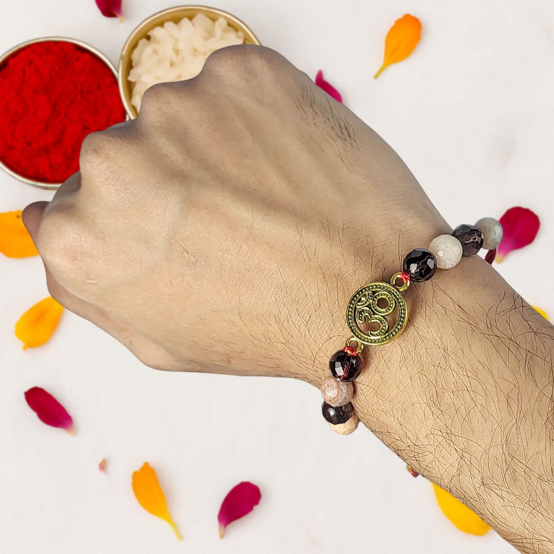 Hand wearing a beaded rakhi with a decorative charm, surrounded by colorful petals on a light background.