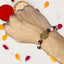 Hand wearing a beaded rakhi with a decorative charm, surrounded by colorful petals on a light background.