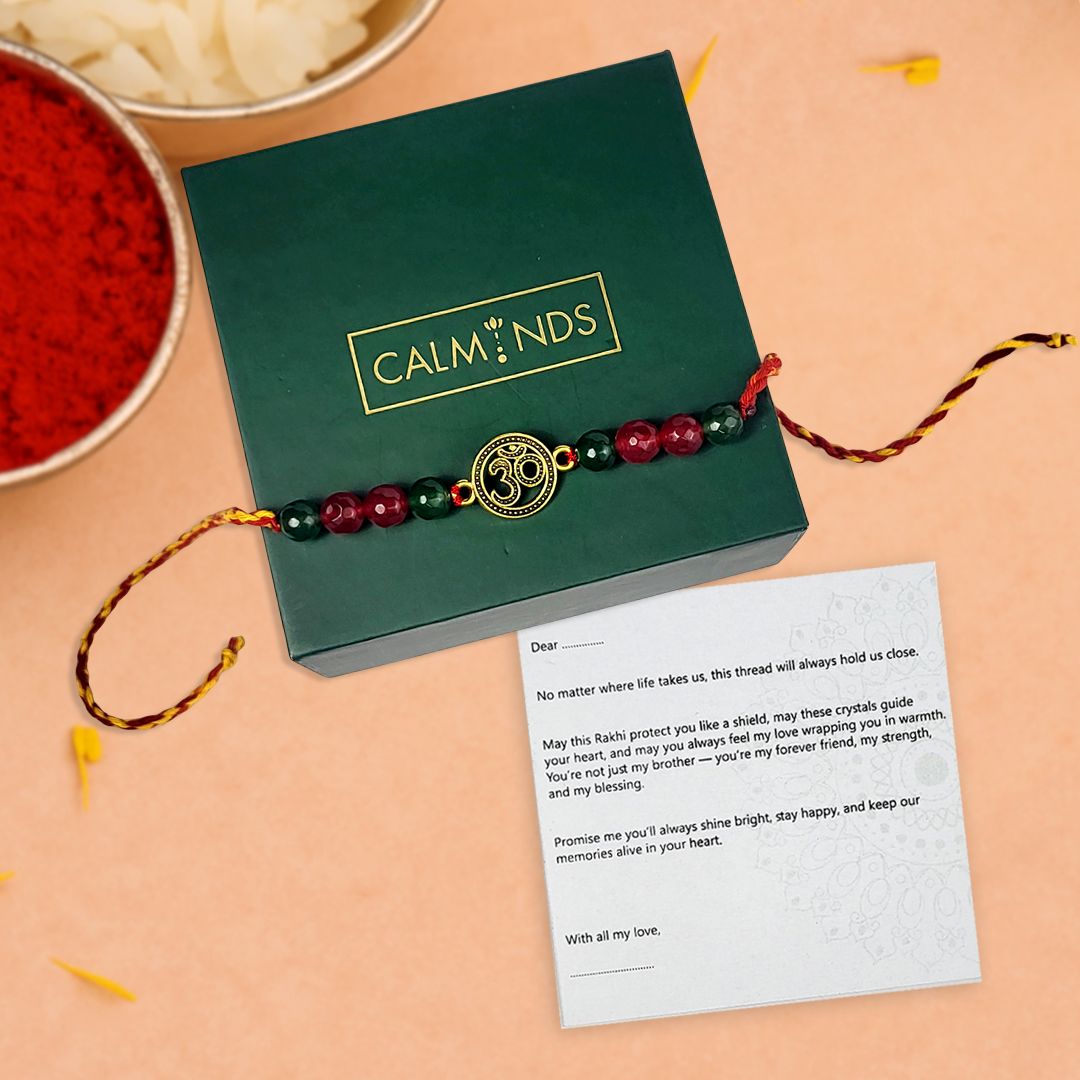 Rakhi with crystals and gold thali on a green 'CALMINDS' box with a card on a peach background.