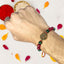 Hand wearing a beaded rakhi with a decorative charm, surrounded by colorful petals on a light background.