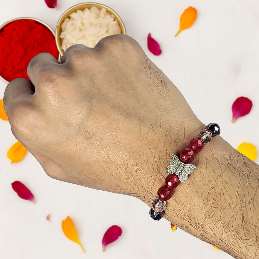 Hand wearing a beaded Rakhi with a butterfly charm, surrounded by colorful petals on a light background