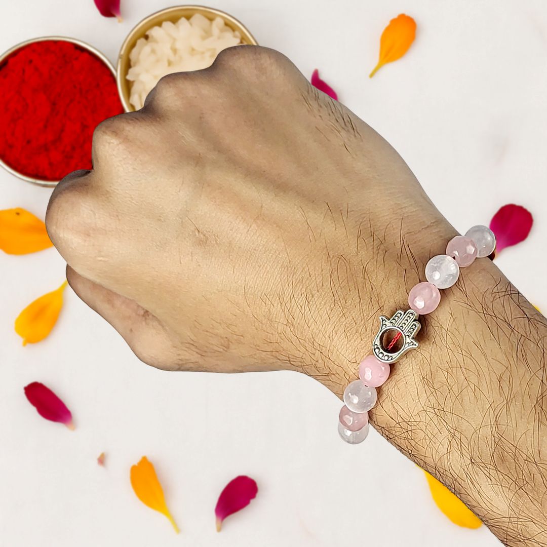 Hand wearing a beaded rakhi with a Hamsa hand charm, surrounded by colorful petals on a light background.