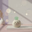 Ceramic figurine with a smiley face and green jade gemstone on a pink surface with blurred lights in the background
