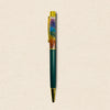 A custom pen with a dark green barrel, gold metal accents, and a clear cap filled with colorful natural resin showing chakra-like patterns, set against a textured beige background.