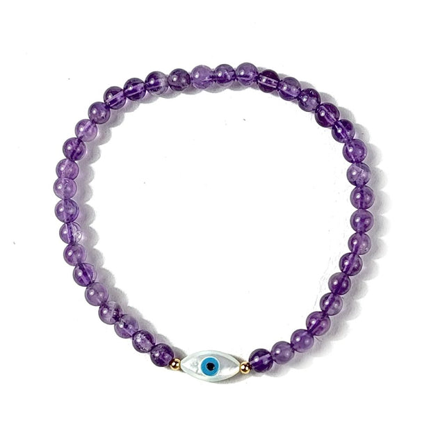Purple beaded bracelet with a gold and silver evil eye charm on a white background