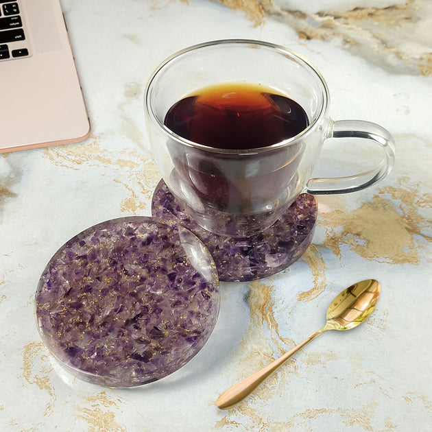 Clear mug with dark liquid on purple amethyst coasters on a marble surface with a laptop.