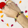 Hand wearing a beaded rakhi with colorful stones, surrounded by flower petals on a light background.