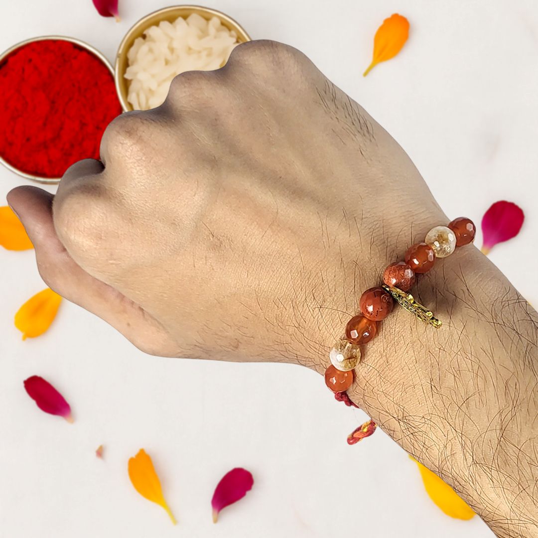 Hand wearing a beaded rakhi with red and gold elements, surrounded by flower petals on a white background.