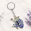 Keychain with peacock design and blue stones on a white background with lavender flowers.