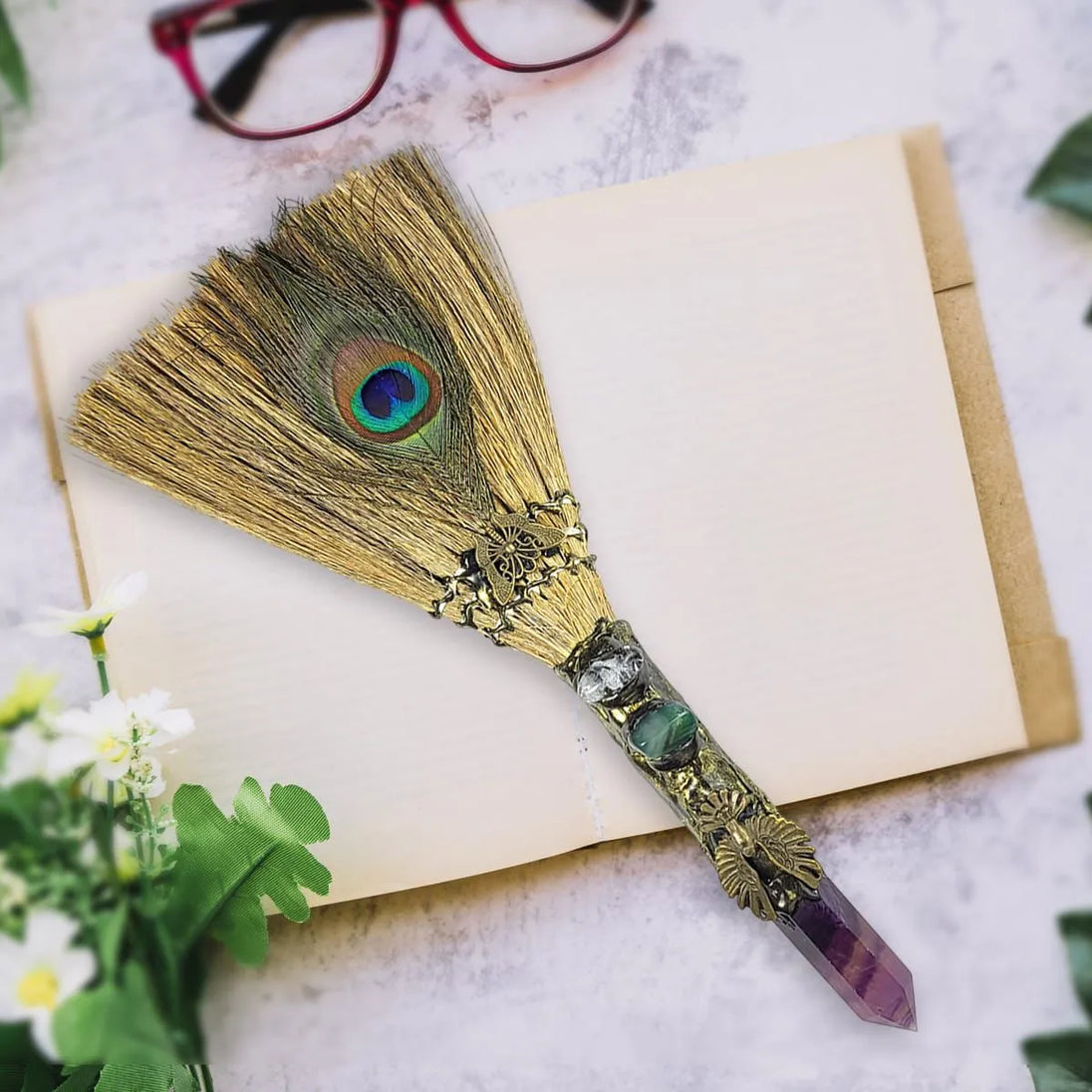 Decorative broom with peacock feathers and crystal handle on a book with glasses and flowers in the background