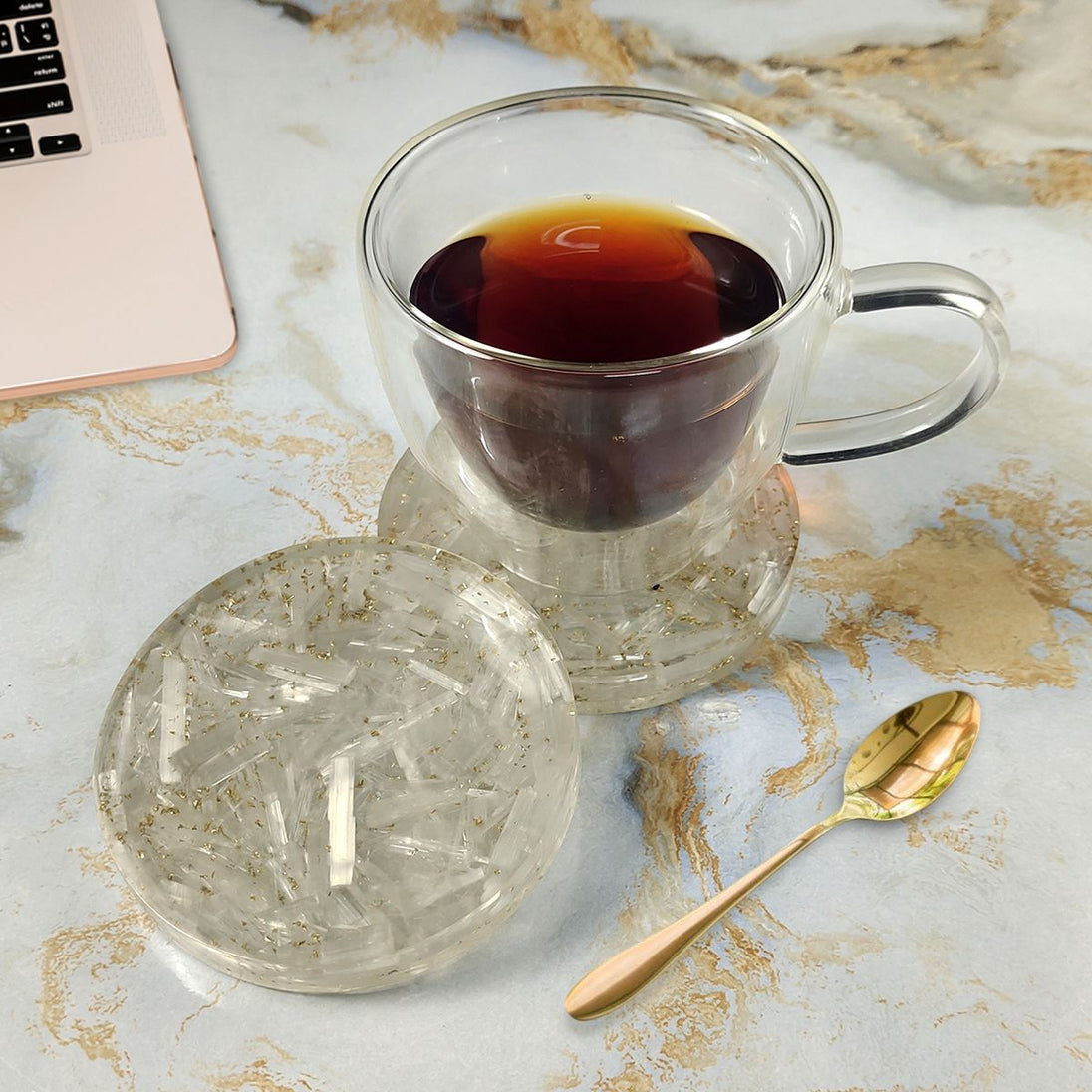 Clear glass mug with dark liquid on a Coaster surface with a laptop and gold spoon.