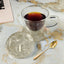 Clear glass mug with dark liquid on a Coaster surface with a laptop and gold spoon.