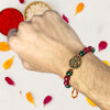 Hand wearing a beaded rakhi with a decorative charm, surrounded by colorful petals on a light background.
