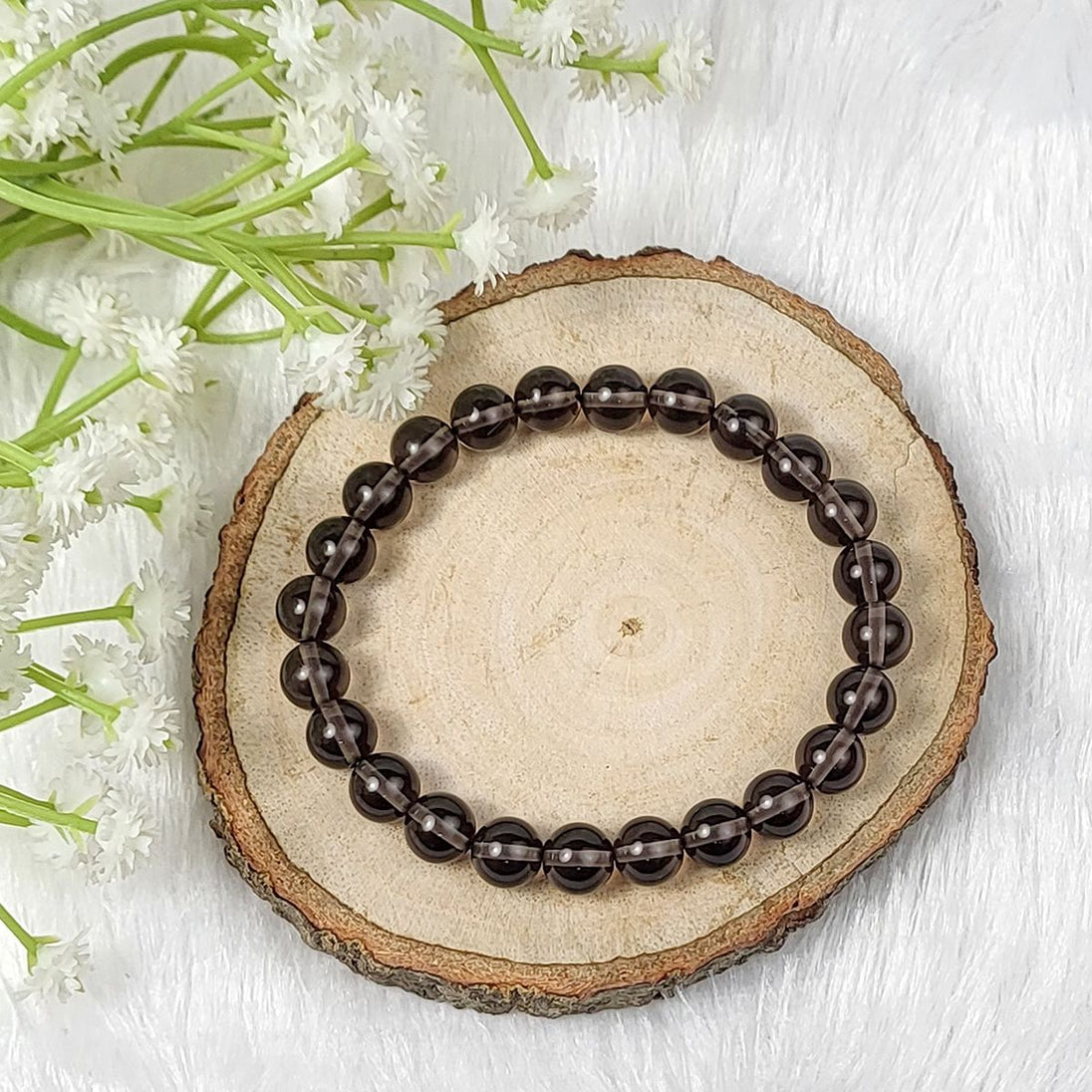 Brown beaded smoky quartz crystal bracelet on a wooden slice with white flowers in the background 