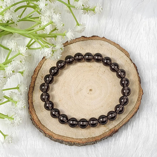 Brown beaded smoky quartz crystal bracelet on a wooden slice with white flowers in the background 