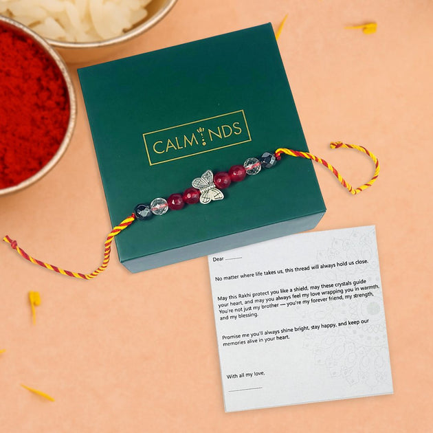 Rakhi with crystals and butterfly charm on a green box labeled 'Calminds' with a card on a peach background.