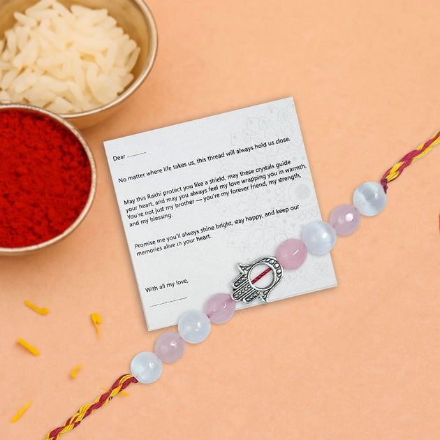 rakhi with pink and yellow crystal beads, a silver hand charm in between on a thankyou card on a peach background