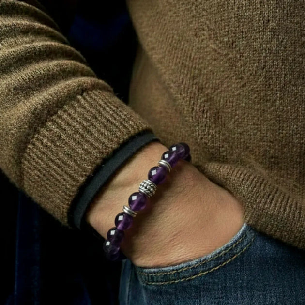 Person wearing a brown sweater and blue jeans with a purple beaded bracelet on a dark background