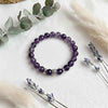 Purple beaded bracelet on a white surface with eucalyptus leaves and lavender flowers.
