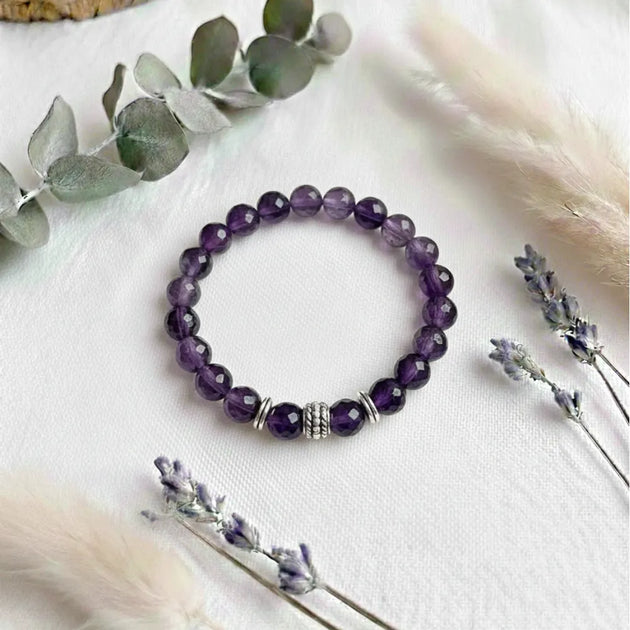 Purple beaded bracelet on a white surface with eucalyptus leaves and lavender flowers.