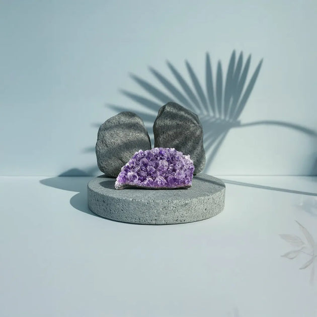 Purple crystal on a gray pedestal with two gray stones against a light blue background