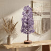 Decorative purple tree on a wooden shelf with a woven wall art piece in the background.