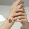 Hand wearing a gold bracelet with a purple crystal on a neutral background