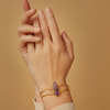 Hand wearing a gold bracelet with a purple crystal on a beige background