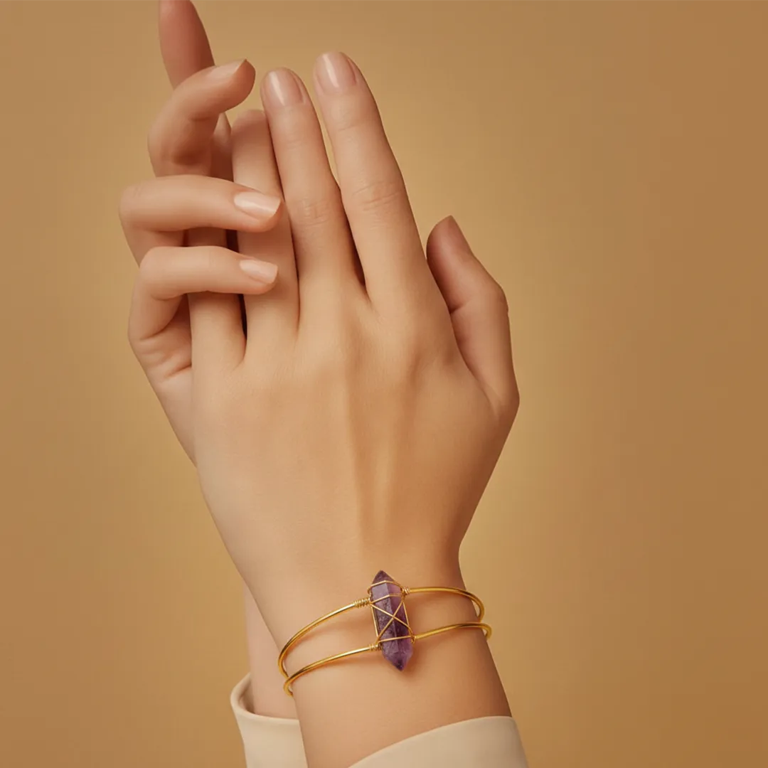 Hand wearing a gold bracelet with a purple crystal on a beige background