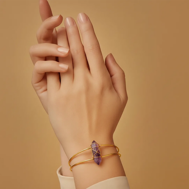 Hand wearing a gold bracelet with a purple crystal on a beige background