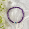 Purple beaded bracelet with a blue evil eye charm on a white fabric background with white flowers.