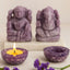 Purple stone statues and candle on a white surface with purple flowers.