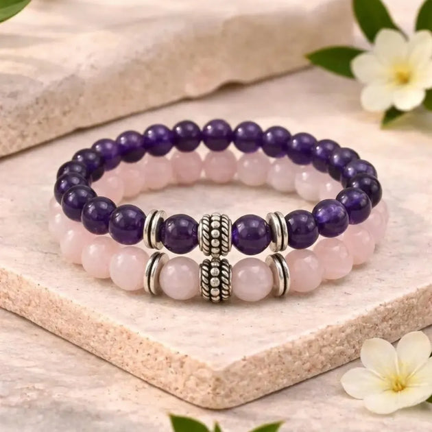 Two beaded bracelets with purple and pink beads on a wooden surface with flowers.