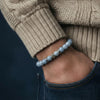 Close-up of a person wearing a beige sweater and blue jeans with a blue beaded bracelet on a dark background