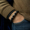 Person wearing a black beaded bracelet on a brown sweater and blue jeans.