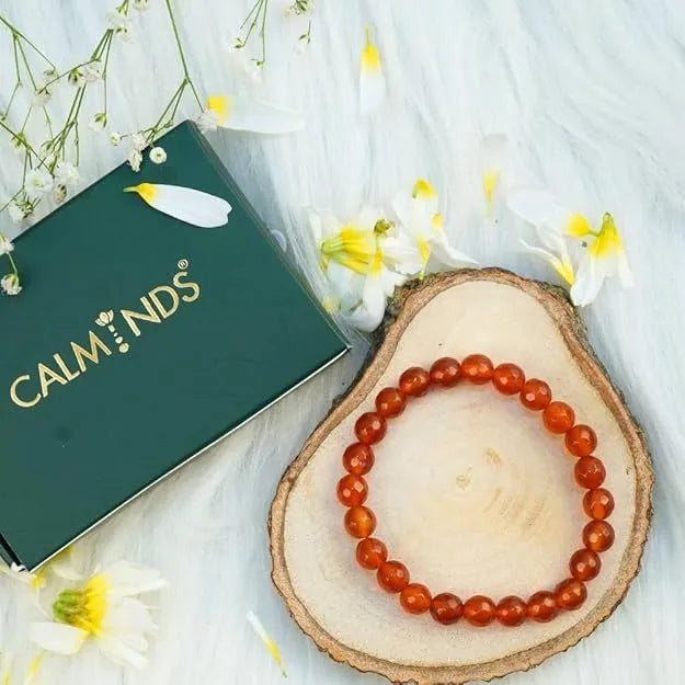 Red beaded bracelet on a wooden slice with a 'CALM NDS' box and flowers in the background