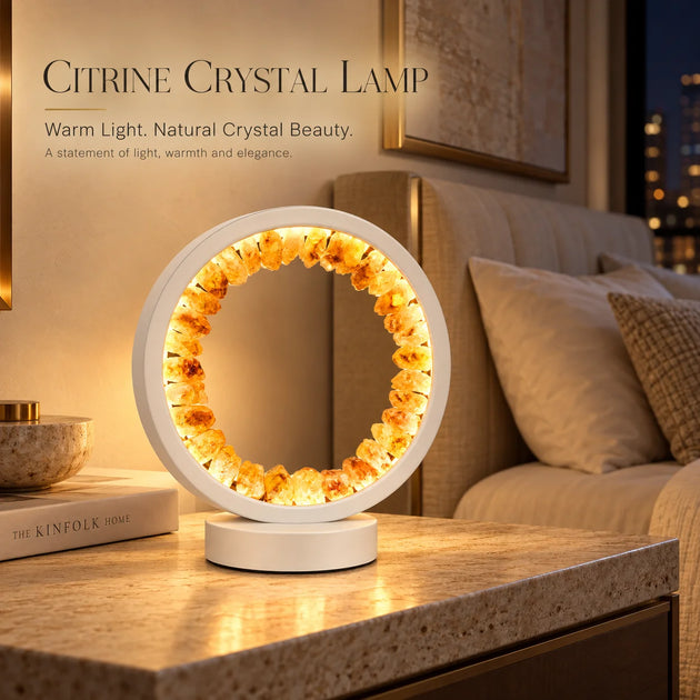 A circular crystal lamp made of warm amber-toned stones on a white base, placed on a stone table in a softly lit bedroom setting. Text at the top reads 'CITRINE CRYSTAL LAMP' with descriptive copy.