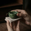 Person holding a small green stone bowl on a cloth with a dark background