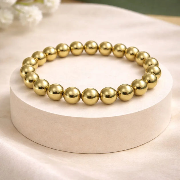 Gold beaded bracelet on a white pedestal with a blurred floral background