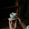 Person holding a small green ceramic cup with a cloth in a dimly lit setting