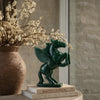 Green winged creature statue on books with a vase of flowers in the background