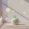 Ceramic figurine with a smiley face and green jade gemstone on a pink surface with blurred lights in the background