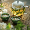 Clear glass teapot with green tea and two small green bowls containing tea and flowers on a wooden surface.