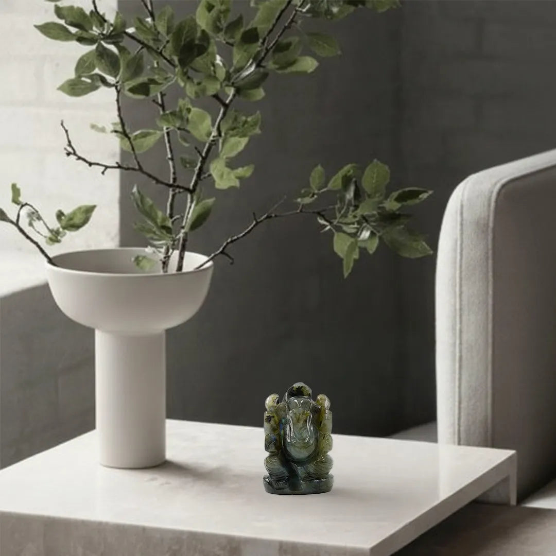 White vase with green branches on a table next to a gray sofa.
