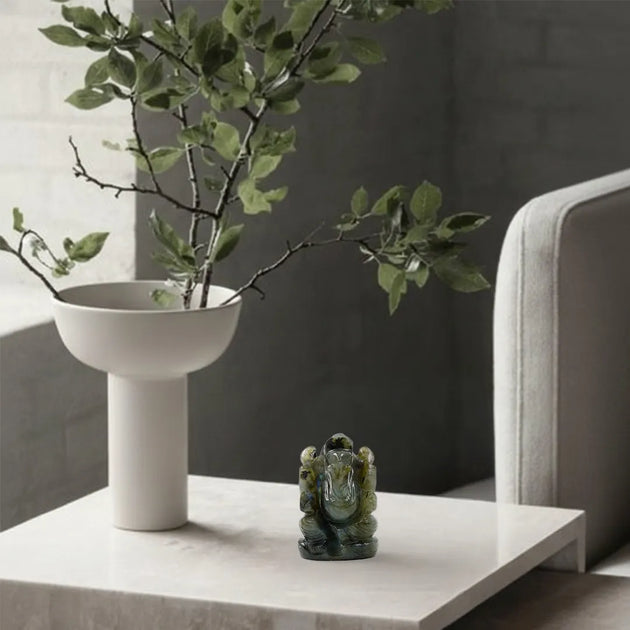 White vase with green branches on a table next to a gray sofa.