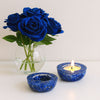 Two blue candle holders with a lit candle and a vase of blue roses on a white surface.