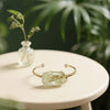 Gold bracelet with a green stone on a white surface with a blurred plant background