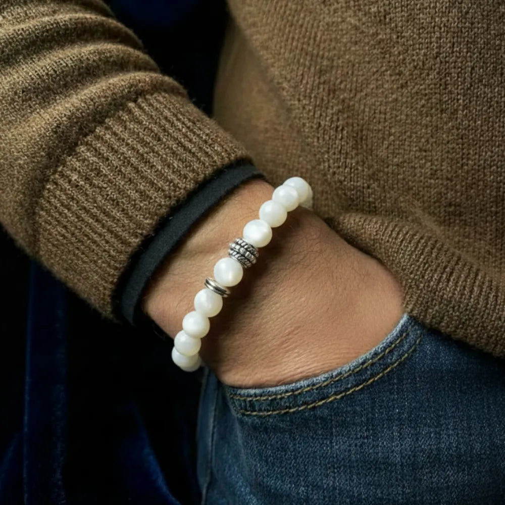 Close-up of a person wearing a white beaded bracelet with a brown sweater and blue jeans.